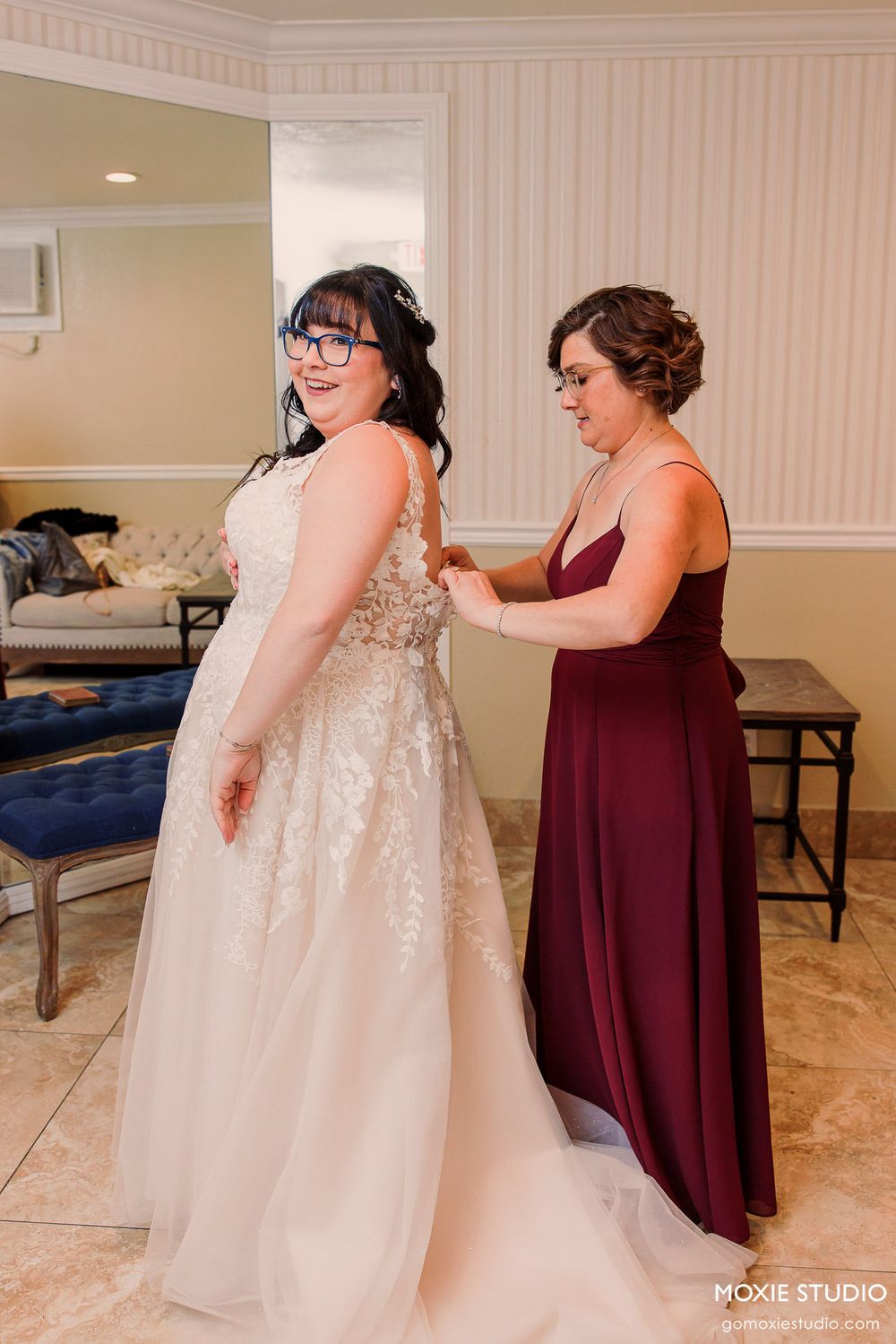 Getting ready scene shows bridesmaids in burgundy dresses helping with a sparkly white wedding gown in a bridal suite.