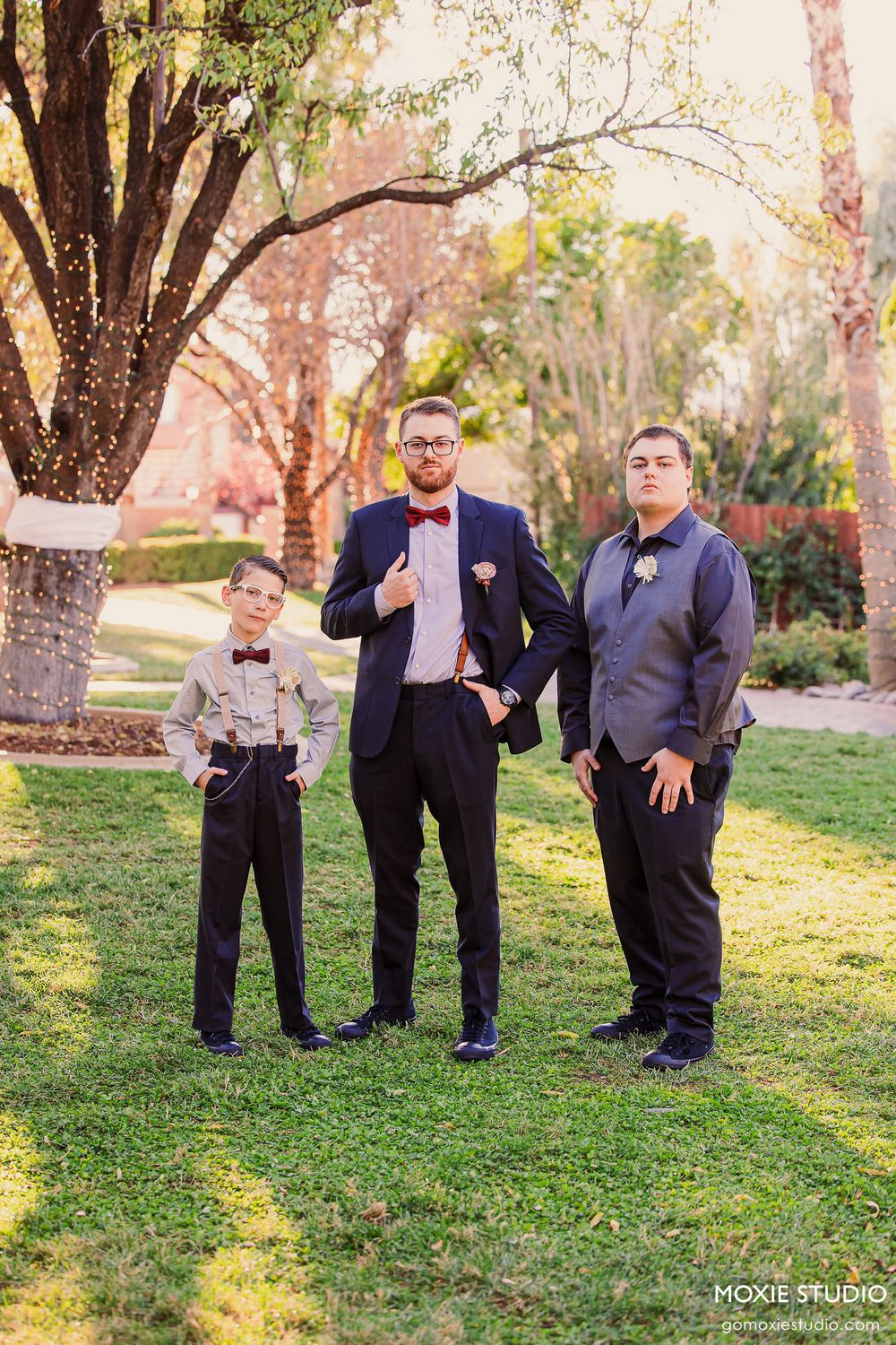 Three groomsmen in formal attire strike playful poses on a grassy lawn during an outdoor wedding photoshoot.