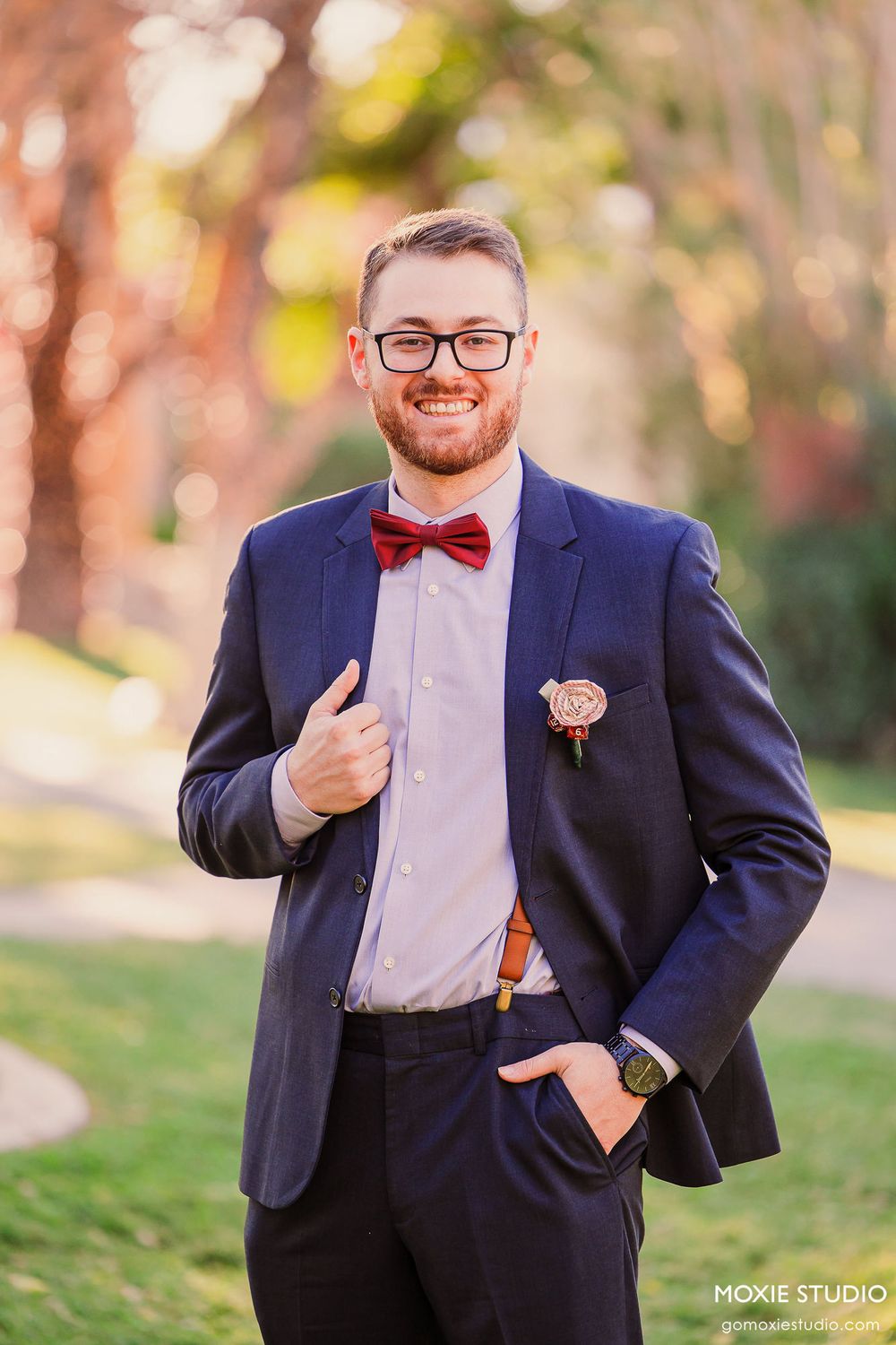 Groomsman in navy suit and red bowtie poses playfully for wedding portraits in outdoor setting.