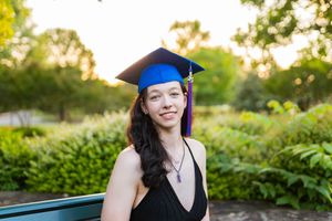Graduate in blue cap and black dress poses outdoors against lush green garden backdrop at sunset.