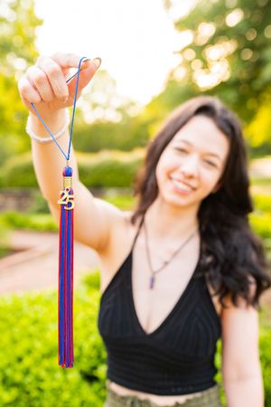 A graduate holds up a purple and blue graduation tassel with decorative charm against a bright outdoor garden setting.