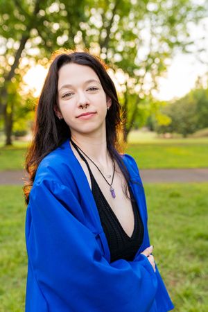A person wearing a bright blue graduation gown and black top poses outdoors in a park during golden hour lighting.