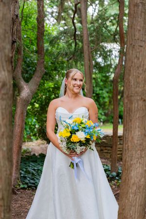Smiling bride in strapless white gown holds yellow and blue bouquet surrounded by trees in a lush garden setting.
