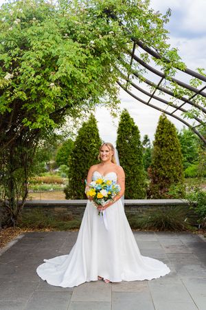 Bride in white off-shoulder gown holding yellow and blue bouquet stands on stone patio beneath garden pergola.