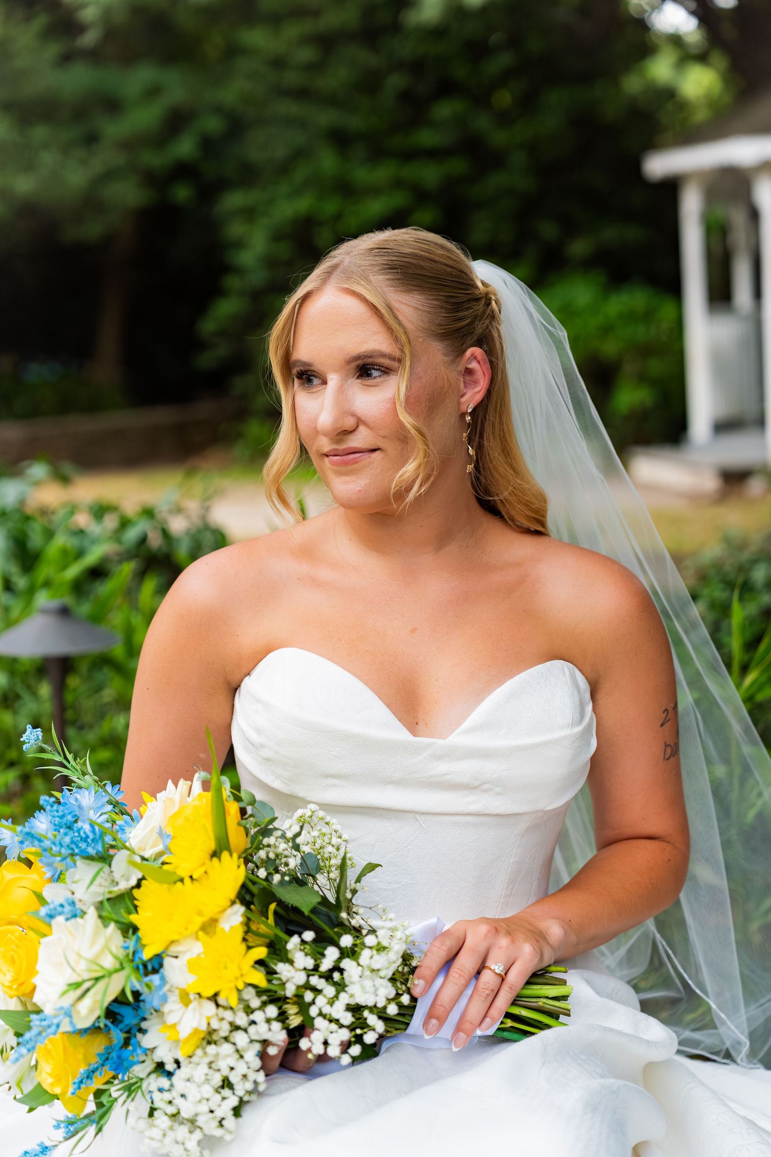 Bride in strapless white gown and veil holding yellow and blue bouquet outdoors at garden wedding venue.