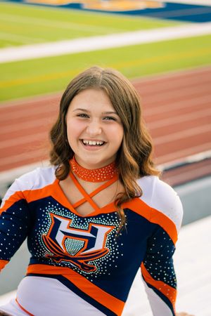 A cheerleader in orange and navy uniform smiles while posing at a sports stadium.