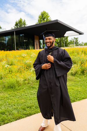 Graduate in black cap and gown stands joyfully on sidewalk with yellow wildflower field and modern building in background.