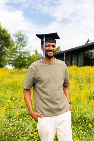 Graduate in cap and gown standing in sunny meadow with yellow wildflowers wearing tan t-shirt and white pants.