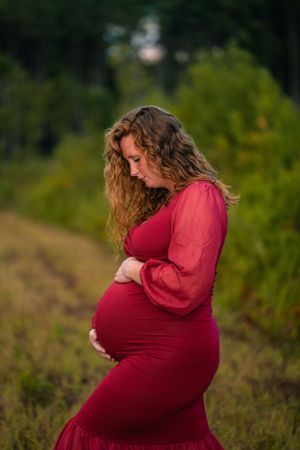 A maternity photo of someone wearing a flowing red dress standing in nature during golden hour.