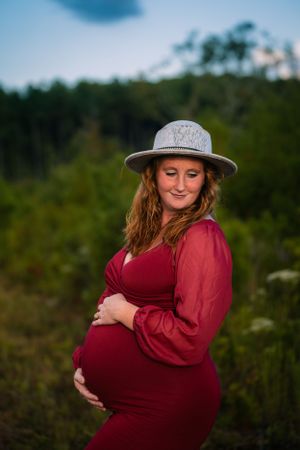 A maternity portrait of a woman in a burgundy dress and hat standing in nature at sunset.
