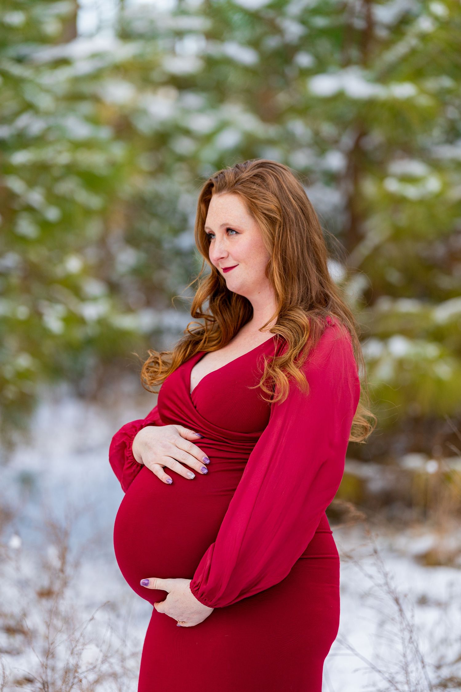 A pregnant person in a red dress poses outdoors in a snowy winter scene among evergreen trees.