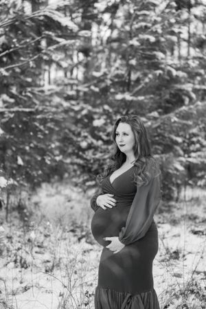 Black and white maternity photo of an expectant mother in a long dress standing in a snowy winter scene.