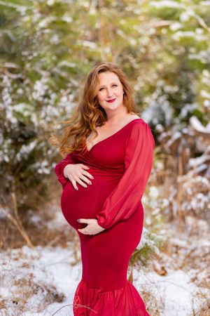 A winter maternity photo features an elegant red dress against a snowy outdoor backdrop with greenery.