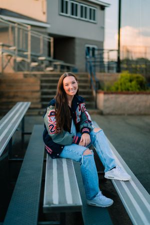 Casual portrait on bleachers wearing a varsity jacket and ripped jeans at sunset.