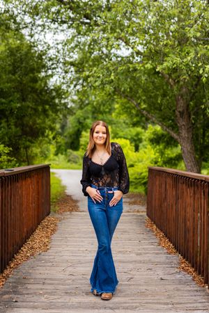 A person in bell-bottom jeans and black top stands on a wooden footbridge surrounded by lush green trees.