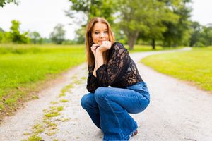 Person crouched on a gravel path wearing blue jeans and black lace top with green grass and trees in background.
