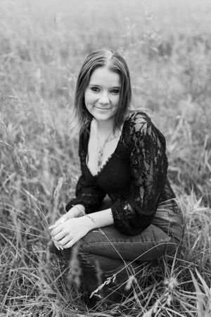 Black and white portrait of young woman in lace top sitting in a field of tall grass.