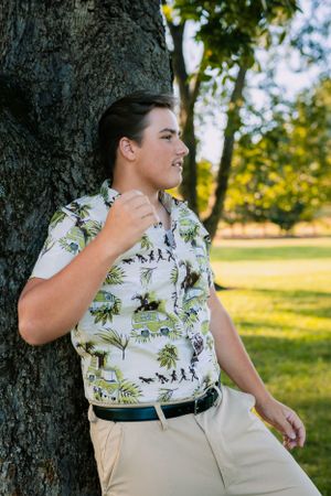 Person in floral print shirt and khakis leaning against large tree trunk.