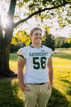 Student athlete wearing white Ravenscroft football jersey number 56 poses by tree at sunset.