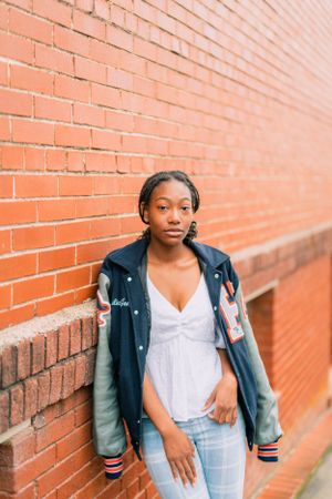 Woman in varsity jacket and jeans posing against orange brick wall.