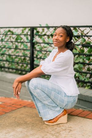 Woman in white peasant top and light jeans crouching casually against leafy backdrop.