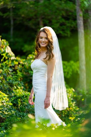 A bride in a white strapless wedding gown and long veil poses in a lush green garden setting.
