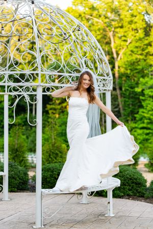 A bride poses gracefully in a white gown under an ornate white metal gazebo.