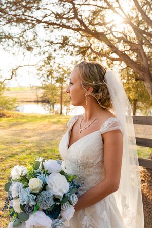 A bride in a white lace gown holds a blue and white bouquet while standing in golden sunlight near a lake.