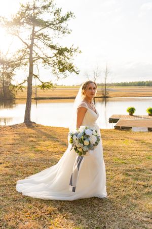 Scenic lake wedding portrait at sunset with elegant white gown and blue floral bouquet near water and pine tree.