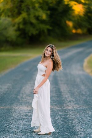 A woman in a flowing white dress poses on a winding country road at sunset.