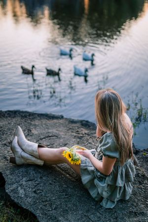 A person in a ruffled dress sits by a pond watching ducks swim at dusk.