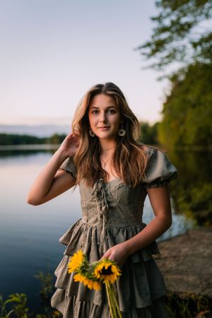 A romantic summer portrait by a lake featuring a ruffled gray dress and sunflowers at sunset.