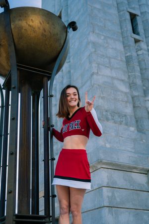 Cheerleader posing in red uniform showing peace sign in front of stone building with bell tower.