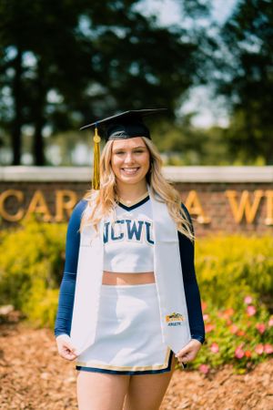 A cheerleader graduate in UCWL uniform poses outdoors near campus signage.