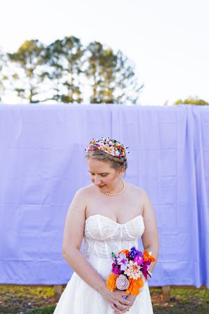Bride in strapless white gown wearing colorful flower crown holds vibrant bouquet outdoors before purple backdrop.