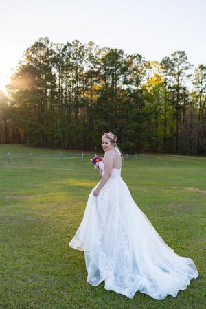 Bride in white ball gown with long train holds colorful bouquet on green lawn at golden hour sunset.