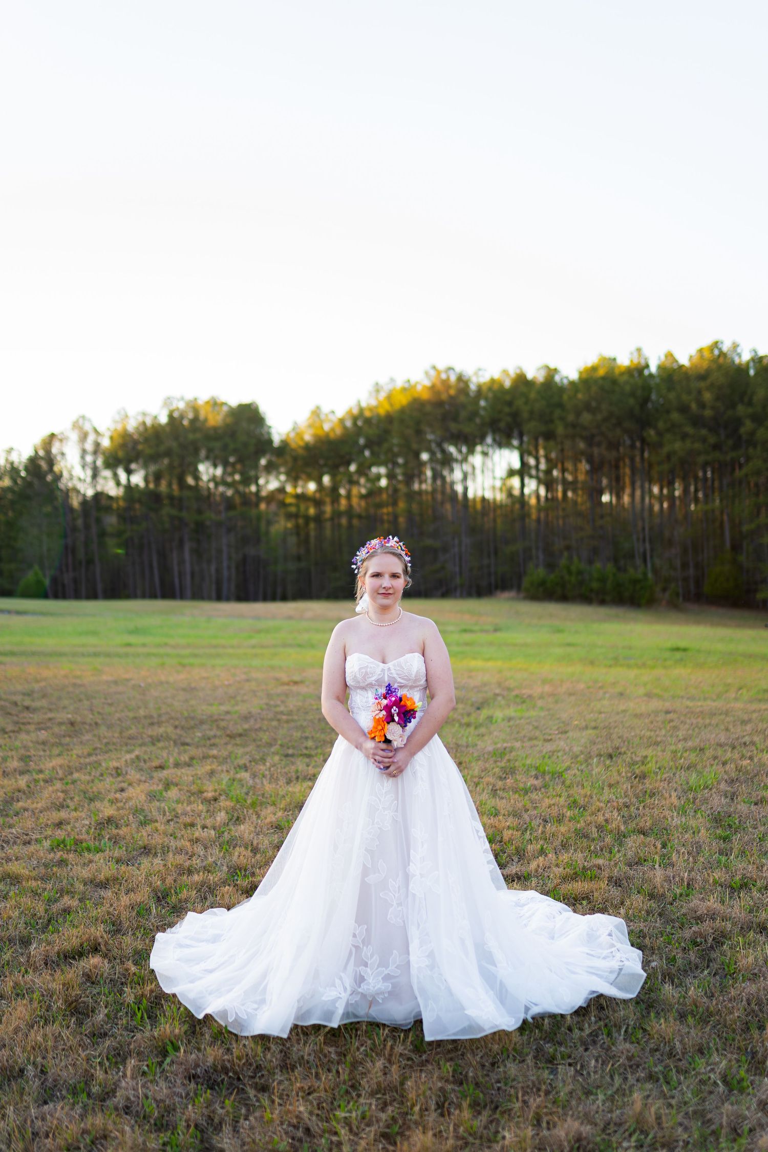 Bride in white ballgown holding colorful bouquet stands in open field with pine trees at golden hour.