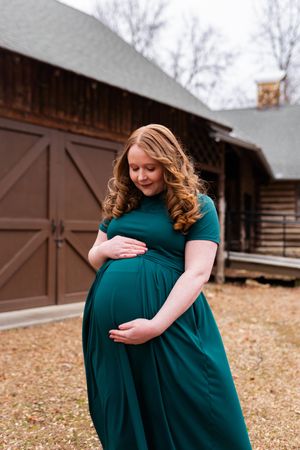A tender maternity photo series in a rustic outdoor setting shows a person in a teal dress cradling their baby bump.