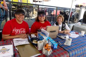 Three people in red Coca-Cola shirts sit at a table with food and drinks at an outdoor event.