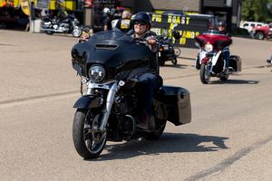 A motorcycle rider cruises on a black touring bike with saddlebags on a sunny street with other bikes visible in background.