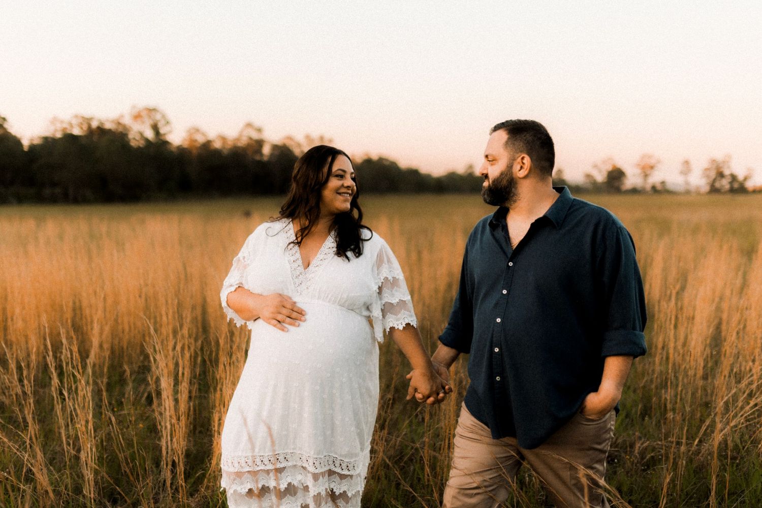 Couple walks together through golden field at sunset during maternity photoshoot.