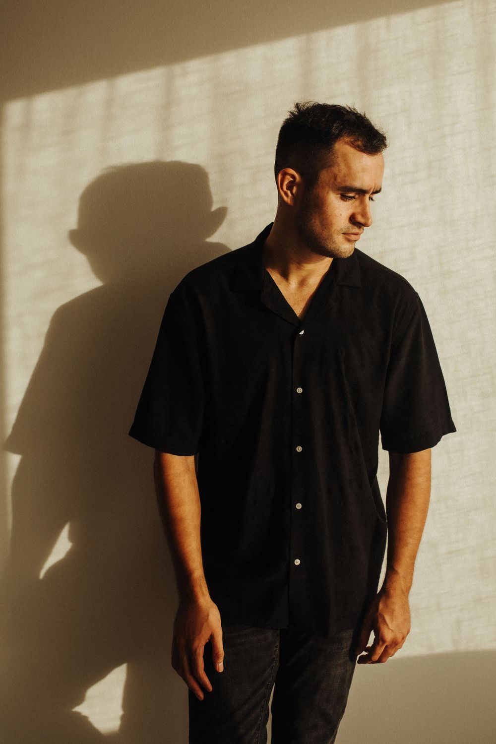 A person in a black shirt stands against a light wall casting dramatic shadows in a minimalist composition.
