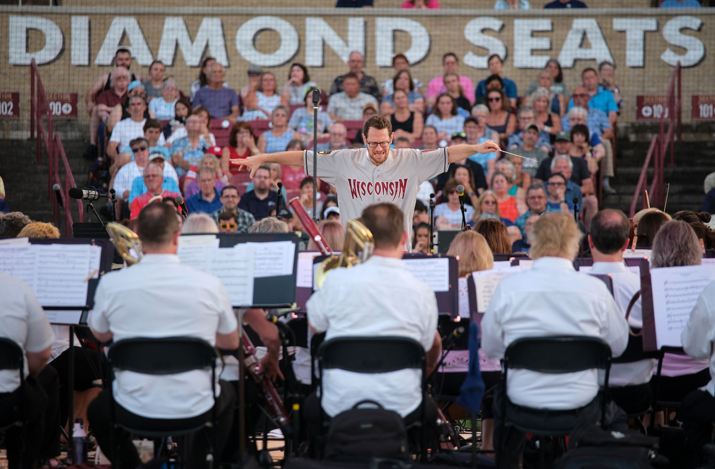 A conductor leads an orchestra while stretching his arms during an outdoor performance.