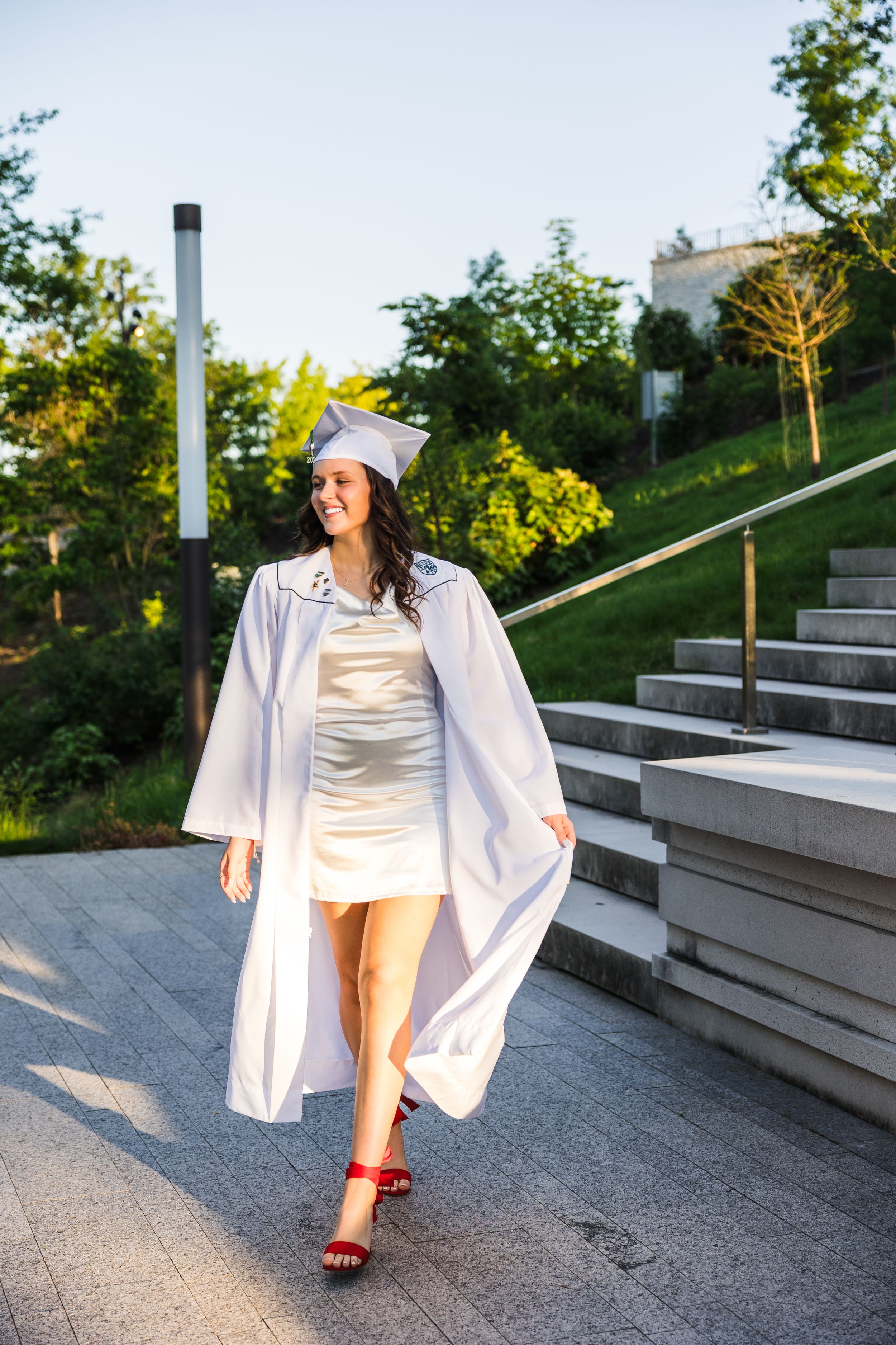 Young woman in white graduation cap and gown walks confidently outdoors on a sunny day near stone steps.