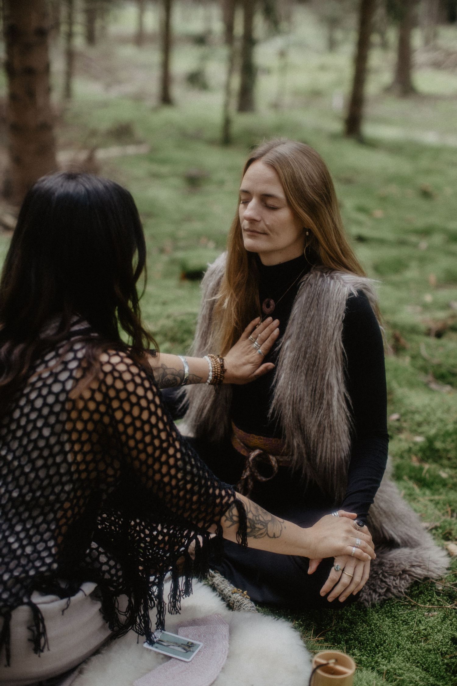 Two friends share a warm embrace while sitting together on grass in a forest setting.