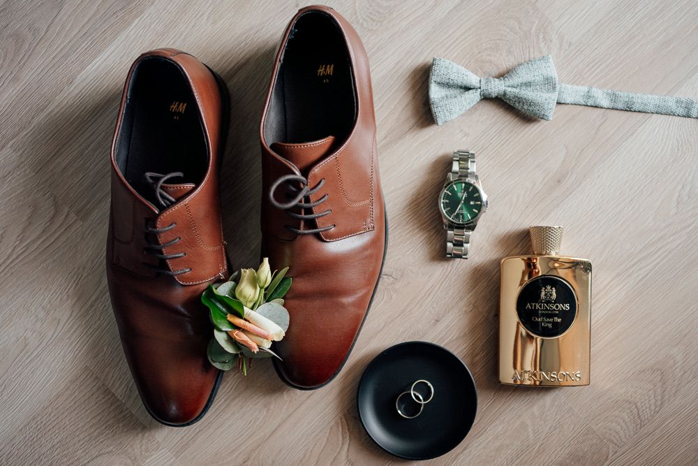 Brown leather dress shoes arranged with bow tie and floral accessories on light wooden floor for wedding styling.