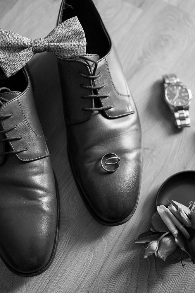 Black leather dress shoes with bow tie and floral details arranged on wooden floor.
