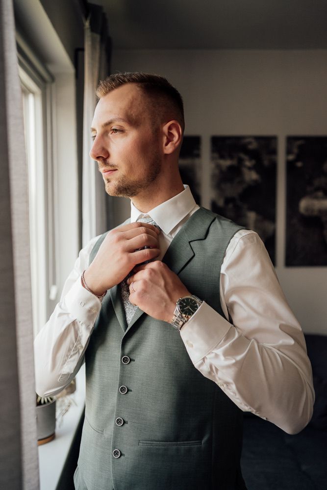 Person wearing a light grey vest and white dress shirt while getting ready near windows.
