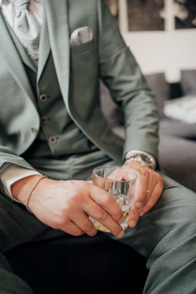 Close-up shots of hands holding a small glass tumbler with a beverage, wearing a sage green suit.