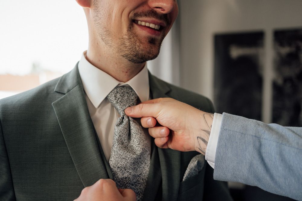 Someone adjusts a silver patterned tie on a dark green suit jacket in an indoor setting.